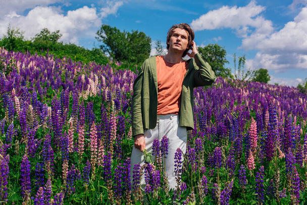 Tall handsome man in a green jacket standing on lupine flowers field holding lupine flowers in hand, enjoing the beauty of nature. Man surrounded by purple and pink lupines. - Zdjęcie, obraz