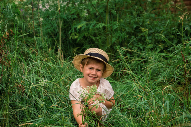 little emotional Girl in a white boho dress with a hat smiling playing in beautiful green thickets of grass. adorable cute girl playing in beautiful thickets in the village. happy childhood - Foto, Imagen