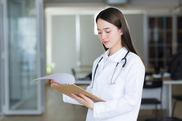 Asiatische Ärztin betrachtet das geöffnete Dokument im Klemmbrett, während sie im Krankenhaus einen weißen Laborkittel und ein Stethoskop trägt. - Foto, Bild