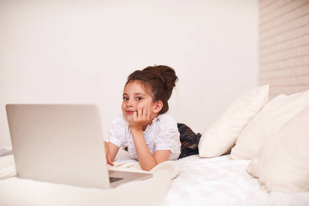little girl is lying on a bed in a room and is using a laptop for training. Watches training videos. E-Education Distance Learning, Home Schooling. Side view. - Foto, afbeelding