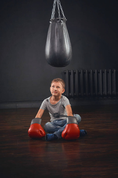 schoolboy boxer, dressed in casual clothes and Boxing gloves, looks at a punching bag and shows a Boxing punch. Isolated on a dark textured background. - Foto, immagini