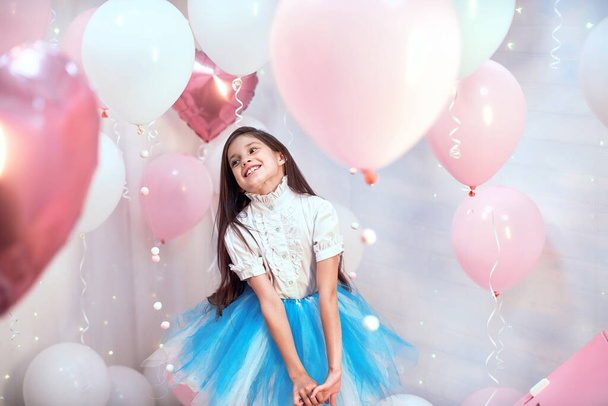 Happy celebration of birthday party with pink helium balloons of charming cute little girl in tulle dress smiling to camera on a blue background . Charming smile, expressing happiness. Place for text. - Photo, image