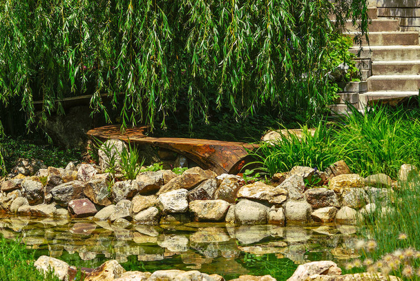 Wooden garden bench in the shape of half a circle by a rocky pond surrounded by greenery - Photo, Image