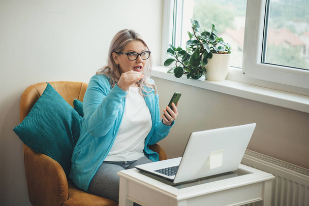 Caucasian woman with blonde hair and eyeglasses explaining something while having an online meeting using a phone and laptop at home - Photo, image