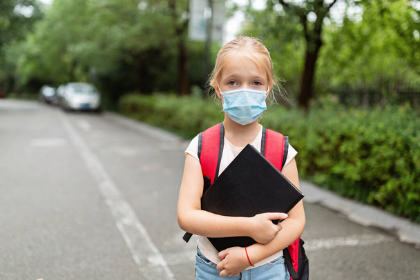 School child wearing face mask during coronavirus pandemic outbreak. Blonde girl going back to school after covid-19 quarantine and lockdown. Kid in medical mask for coronavirus prevention. New normal. High quality photo - Foto, Bild