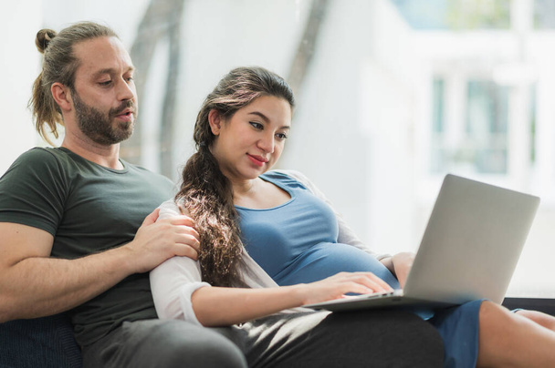 Baard man en zijn zwangere vrouw kijken film op laptop zich gelukkig voelen terwijl ze op de bank in huis zitten. Gezinsconcept. - Foto, afbeelding