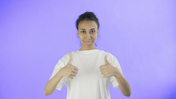 Beautiful young girl in a white T-shirt shows thumb up with both hands on Violet background - Photo, Image