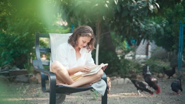 A young woman sits in a chair in the garden and reads a book - Filmati, video