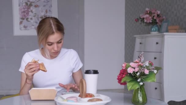 Genç bir sarışın kadın haberleri Smartphone 'da okuyor. Hafif bir kafede bir sandalyede oturuyor, bir kadın kahve içiyor ve öğle yemeği molasında sandviç yiyor. - Video, Çekim