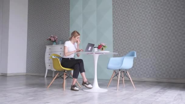 Genç sarışın bir kadın bilgisayarda çalışıyor. Parlak bir kafedeki bir masada, bir sandalyede oturuyor. - Video, Çekim