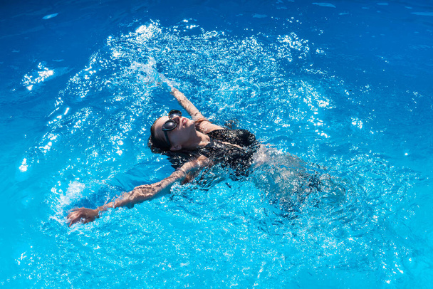 Meisje in zwart badpak en zonnebril zwemt op haar rug in helder water in het zwembad. Mooie vrouw in een zwembad. blauw water van het zwembad. borstslag, kruipen, vlinder - Foto, afbeelding