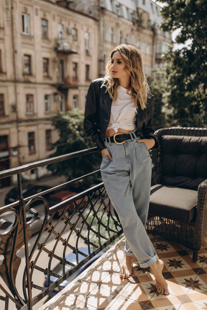 Stylish fashionable blonde woman with smoky eye makeup, in jeans, white t-shirt and black leather jacket on the balcony in the city. Spring autumn fashion concept. Soft selective focus. - Photo, Image