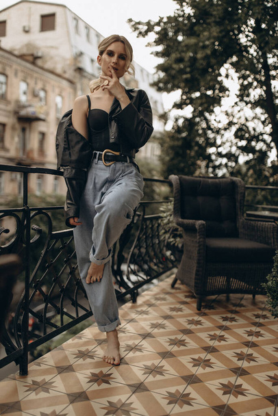 Stilvolle blonde Frau mit rauchigen Augen, in Jeans, Dessous und schwarzer Lederjacke auf dem Balkon der Stadt. Frühling Herbst Modekonzept. Weiche selektive Fokussierung. - Foto, Bild