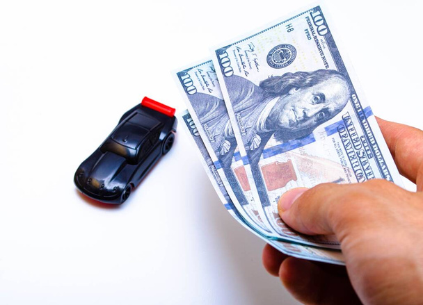 Мужская рука держит долларовые купюры на фоне черного спортивного автомобиля на белом фоне. Покупка машины, продажа машины - Фото, изображение