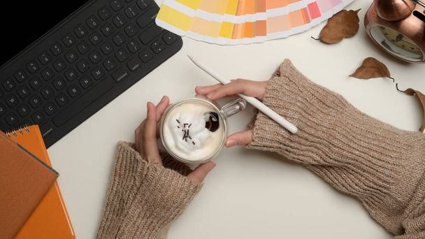 Overhead-Aufnahme weiblicher Designerhände, die einen Becher mit Getränken auf einem Arbeitstisch mit Vorräten im Home Office-Raum halten  - Foto, Bild