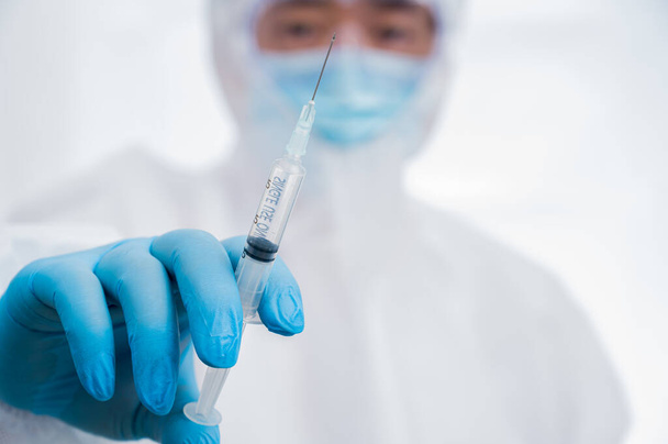 A male scientist wearing blue gloves and a protective suit holding a Covid-19 vaccine. - Фото, изображение