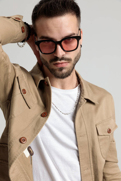 fresco giovane uomo non rasato in giacca indossa occhiali da sole tenendo la mano dietro il collo in una posa di moda su sfondo grigio - Foto, immagini