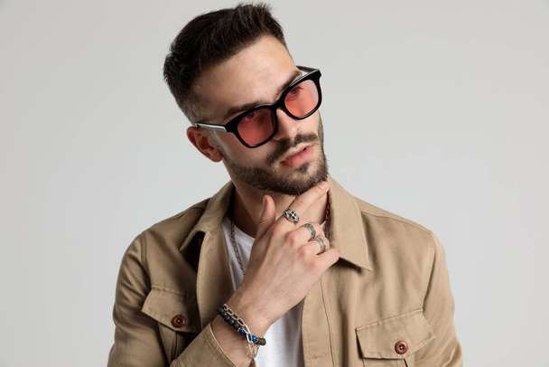 thoughtful unshaved young man in jacket wearing sunglasses, touching chin and thinking, looking to side and posing on grey background - Photo, Image