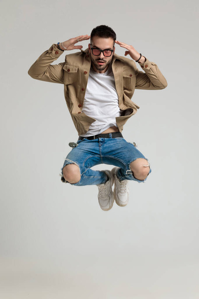 cool young casual guy in jacket wearing sunglasses jumping and pointing fingers to head, posing on grey background - Photo, Image