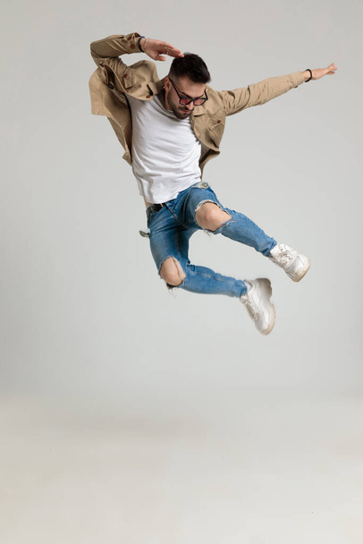 cool young man in jacket wearing sunglasses, looking down, holding hands in the air and jumping on grey background - Foto, Bild