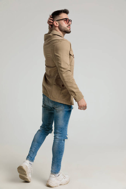back view of young casual guy wearing glasses, looking to side and scratching head, thinking, walking on grey background - Foto, Imagen