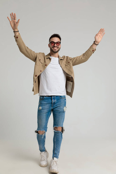 glimlachende jonge casual man in jas met zonnebril, omhoog kijken en hand in hand in de lucht juichen overwinning en trots lopen op grijze achtergrond - Foto, afbeelding