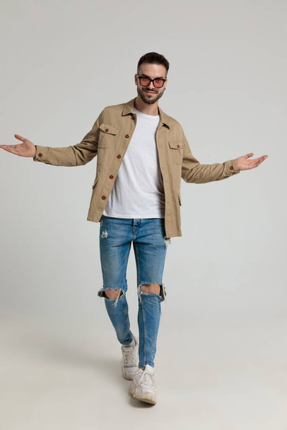 young casual unshaved man in jacket wearing sunglasses, opening arms and welcoming, walking and posing on grey background - Foto, Imagem