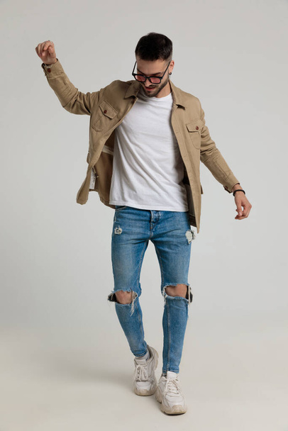 happy young casual guy in jacket wearing sunglasses, looking down, holding hands in the air and posing, walking on grey background - Photo, Image