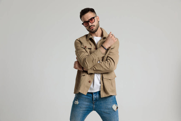 serious cool young man in jacket wearing sunglasses and holding elbows in a fashion pose, standing on grey background - Fotografie, Obrázek