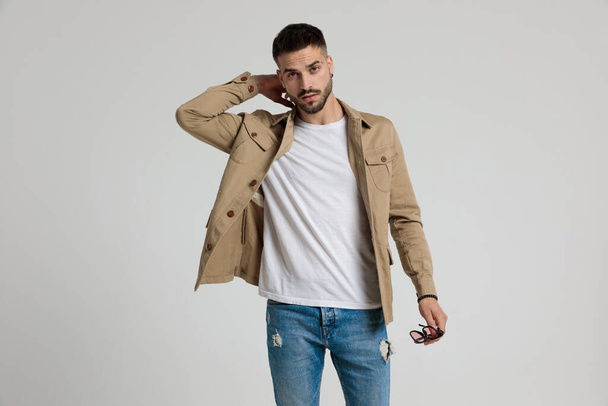 sexy young casual man in jacket holding sunglasses, holding hand behind neck in a fashion pose, standing and posing on grey background - Photo, image