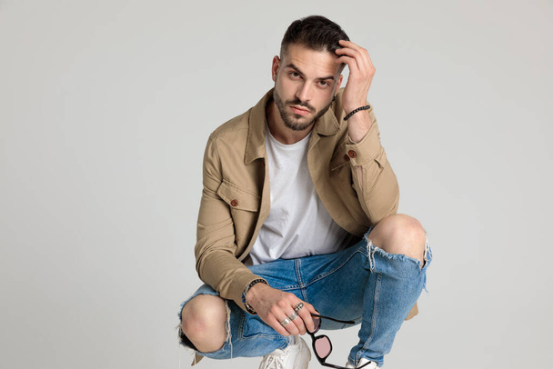 young unshaved model in jacket holding sunglasses, running fingers through hair, holding elbows on knees and crouching on grey background - Fotografie, Obrázek