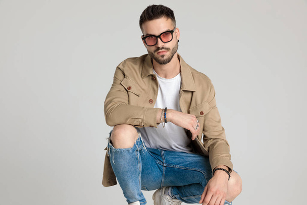 young unshaved casual model in jacket wearing sunglasses, holding elbows on knees and crouching on grey background - Foto, immagini