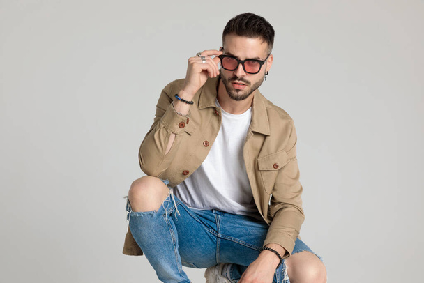 confident young unshaved man in jacket fixing sunglasses, holding elbows on knees and crouching on grey background - Foto, Imagen