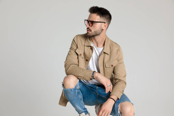 sexy young casual model wearing beige jacket and sunglasses, looking to side, holding elbows on knees, bending the knee and crouching on grey background - Photo, Image