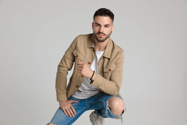 handsome young casual guy in jacket holding elbow on knee, bending the knee and crouching on grey background - Фото, зображення