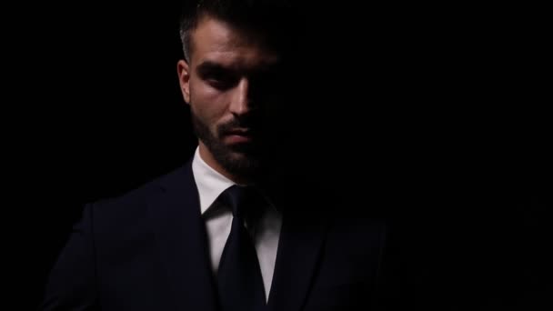 attraktives junges Mode-Model im Anzug, im Schattenlicht stehend und Krawatte fixiert, posierend und Arme verschränkend auf dunklem Hintergrund - Filmmaterial, Video