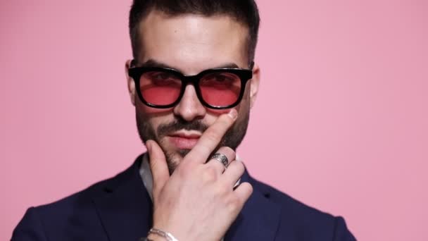 primo piano di un uomo d'affari sexy cool con gli occhiali da sole, toccandosi la barba, facendo un gesto di silenzio, girandosi e lasciando su sfondo rosa - Filmati, video