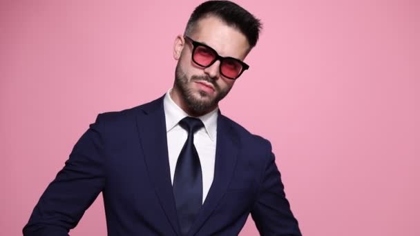 жорсткий модний бізнесмен фіксує краватку, одягаючи сонцезахисні окуляри, позуючи зі стилем, набиваючи щоки, стирчать язиком і обманюють навколо на рожевому фоні
 - Кадри, відео