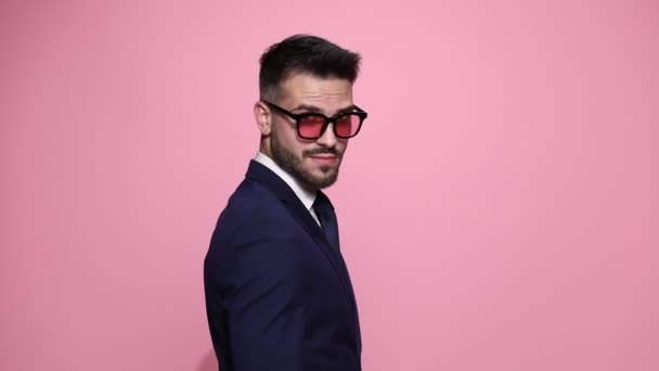 attractive businessman standing in rear view, slowly turning with his face at the camera, arranging his jacket and his tie, looking away, fixing his sunglasses, gesturing a come here sign on pink background - Footage, Video