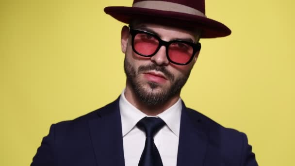 confident young businessman in suit wearing hat and sunglasses, moving head in both sides, looking, snapping fingers and walking away on yellow background - Footage, Video