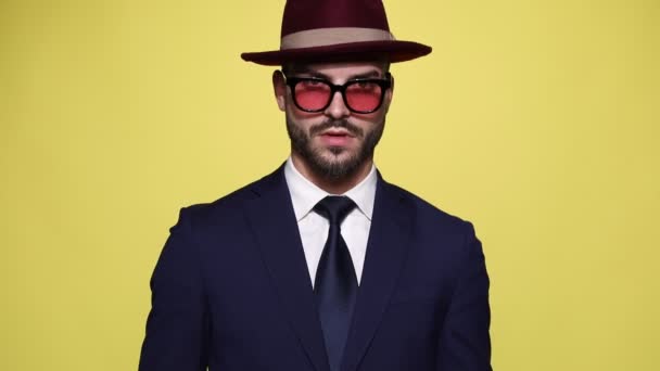 aantrekkelijke jonge zakenman in pak dragen hoed en zonnebril, kijken naar beneden en omhoog, bewegend hoofd en vaststelling van pak en bril, bewegen in een zijaanzicht positie en lopen weg op gele achtergrond - Video