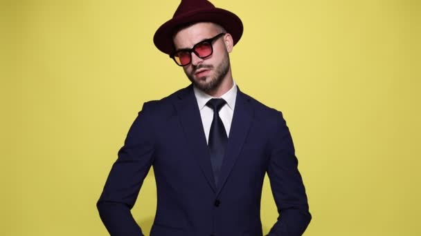 sexy hombre de negocios en traje con sombrero y gafas de sol moviéndose y sonriendo, arreglando el traje y mirando hacia abajo, frotando las palmas y el traje de fijación, alejándose y posando en una posición de vista trasera sobre fondo amarillo
 - Metraje, vídeo