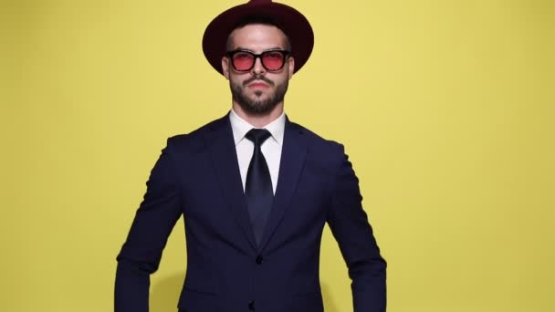 elegant young businessman in suit wearing sunglasses and hat, holding hands on hips and standing on yellow background - Footage, Video