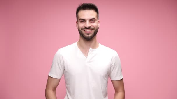 young casual man standing against pink background, looking at camera and smiling, shrugging his shoulders, without a clue, looking aside on pink background - Footage, Video