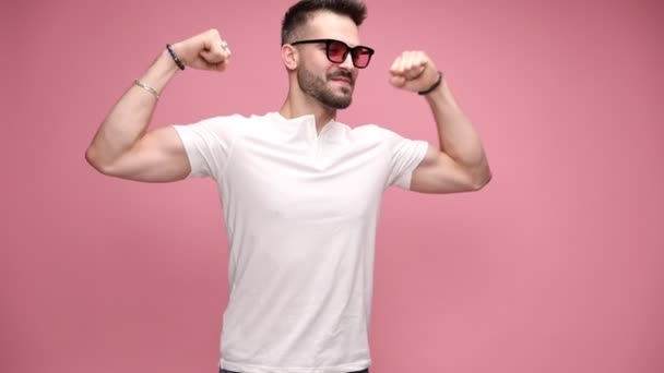 сексуальный случайный мужчина разминает руки, показывает свои мускулы, надевает солнечные очки, целует бицепсы, проявляет мужественность и силу, но просто дурачится и рвется в смех на розовом фоне - Кадры, видео