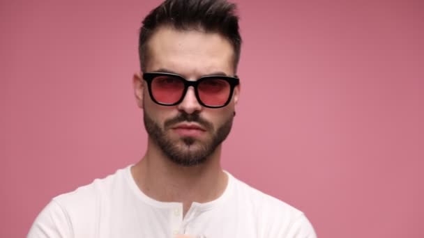 close up of a serious casual man emodying a tough attitude, fixing his sunglasses, being surprised and bursting to laugh, looking aside and covering his eyes on pink background - Footage, Video