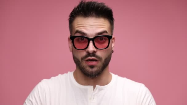 közeli egy szexi alkalmi férfi visel napszemüveg, belenéz a kamerába, meglepett, tegye a kezét a fejére, emelje fel a szemüvegét, és fedezze a szemét, kukucskál ujjai között rózsaszín háttér - Felvétel, videó
