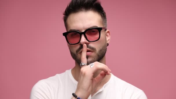 close up of a cool casual man gesturing a shut up sign, fixing his sunglasses, touching his neck and turning his back, being seductive on pink background - Footage, Video