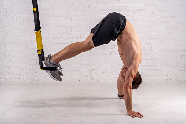 Muscular young man doing exercises with trx straps, near a white brick wall. Active lifestyle, bodybuilding. - Fotó, kép