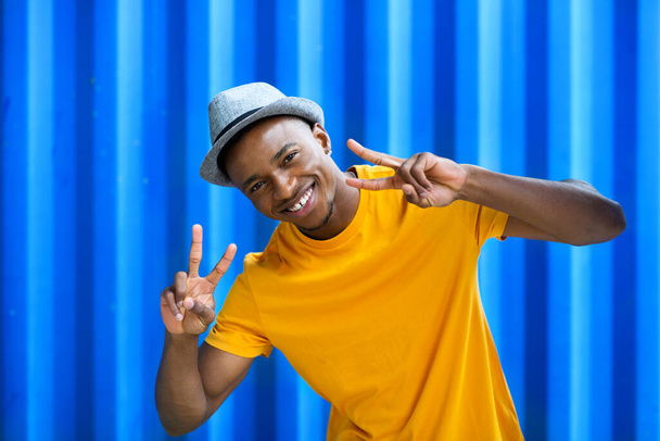 Cheerful young black man standing against blue background, peace gesture concept. - Foto, imagen